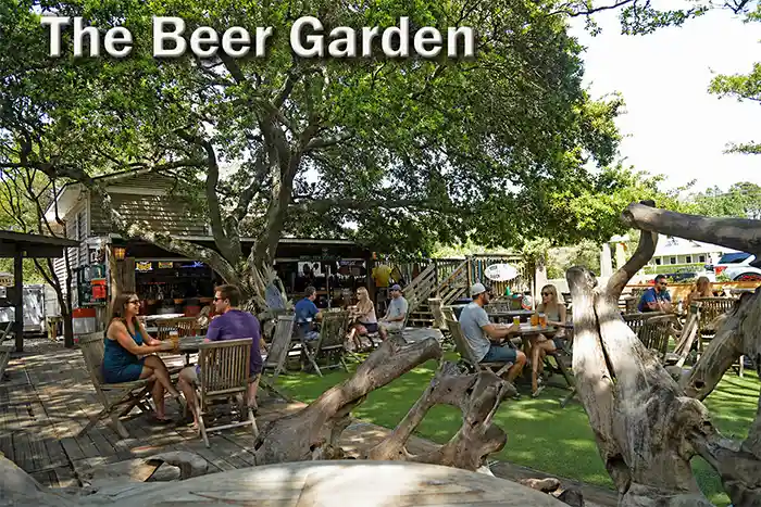 Outdoor beer garden in Corolla, North Carolina with people sitting at wooden tables under large shade trees enjoying drinks in a relaxed, rustic setting.
