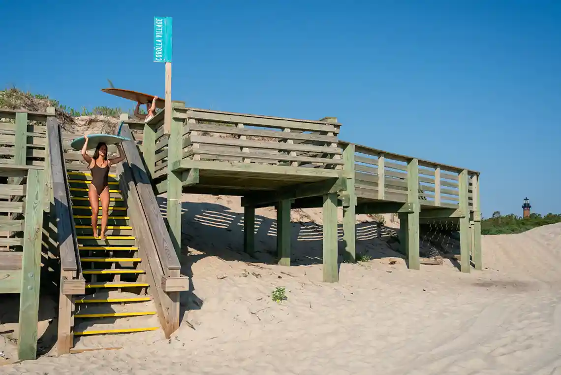 Corolla NC beach access boardwalk with surfers carrying boards over the dunes and Currituck Beach Lighthouse in the distance