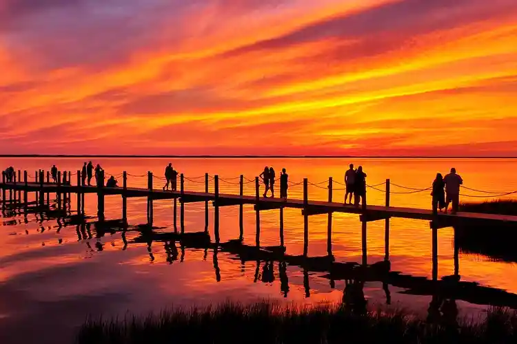 Vibrant sunset over the Duck, North Carolina boardwalk with silhouettes of people walking along the pier and reflections glowing across the calm sound waters.