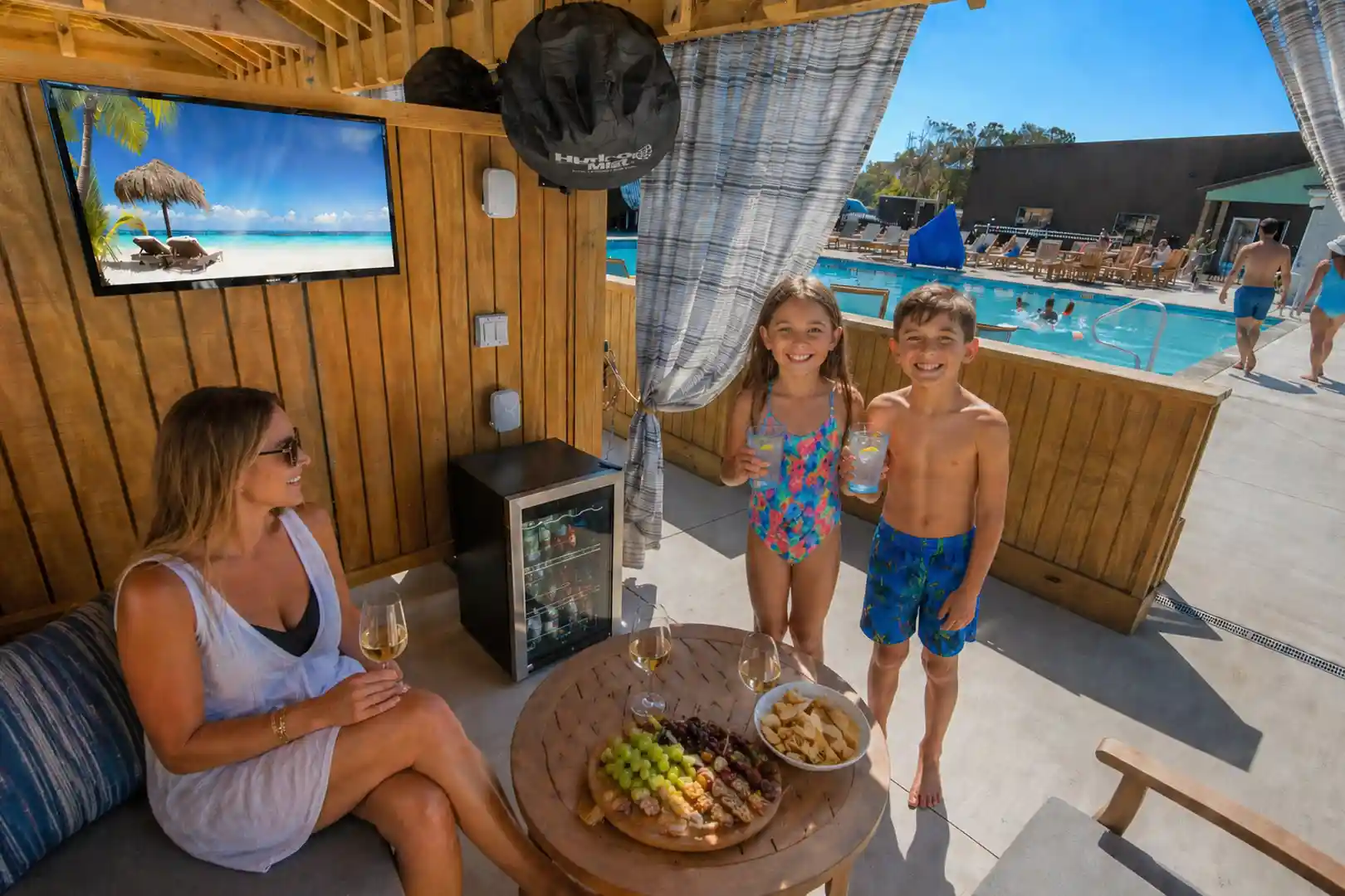 Family enjoying Sonoma’s Hideaway poolside cabana in Corolla NC with snacks, smiling kids, and pool view