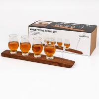 Whisky stave flight set with four Glencairn glasses filled with whisky and glass pipette displayed on a reclaimed oak barrel stave in front of product packaging.