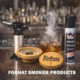 Foghat cocktail smoker kit with wooden smoker lid, culinary torch, butane refill, and smoked whiskey in a rocks glass on a bar counter with smoke rising.