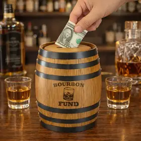 Mini oak barrel piggy bank with engraved Bourbon Fund design on whiskey bar with glasses and dollar bill being inserted