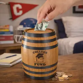 College Drinking Fund mini oak barrel piggy bank sitting on a dorm room desk with a hand inserting a dollar bill and loose coins around the base.