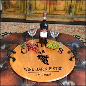 'Grapes' Barrel Head Serving Tray 21-inch round reclaimed oak barrel head serving tray with wrought iron handles displaying wine bottle, glasses, and grapes in a rustic wine bar setting.