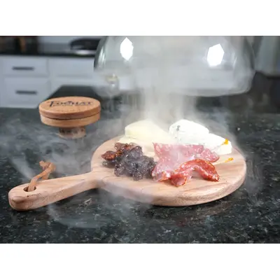 Glass smoking cloche dome with round wooden cutting board and handle with smolker in the background and smoke rising from the food