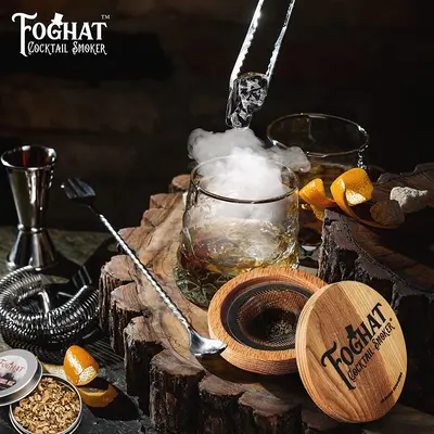 Smoked Old Fashioned cocktail on rustic bar surface with Foghat oak smoker lid and bourbon garnish.