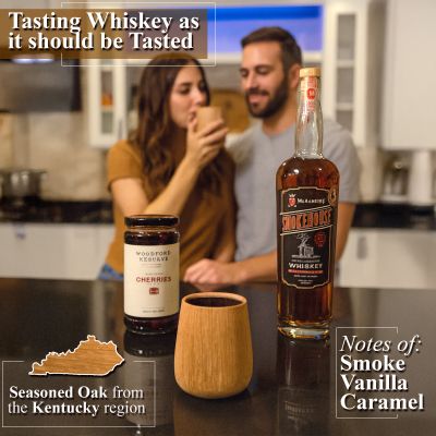 A woman and man in a modern kitchen tasting whiskey from a wooden tumbler, with a bottle of Smokehouse whiskey, Woodford Reserve cherries, and a charred oak tumbler displayed on the counter.