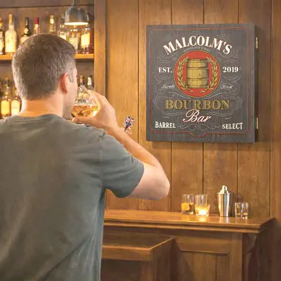 Man drinking whiskey while playing darts in a home bar featuring a personalized Bourbon Bar dartboard cabinet with custom name design, perfect for man cave or whiskey bar decor.